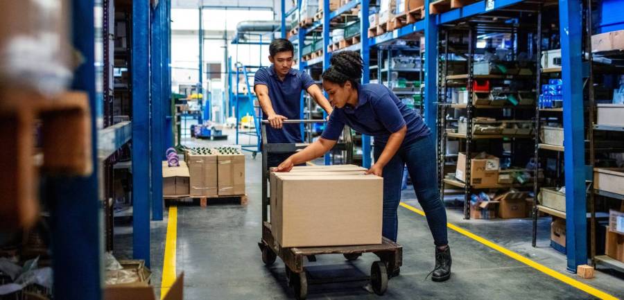 Two warehouse workers handling a heavy box on a cart in an industrial storage facility.